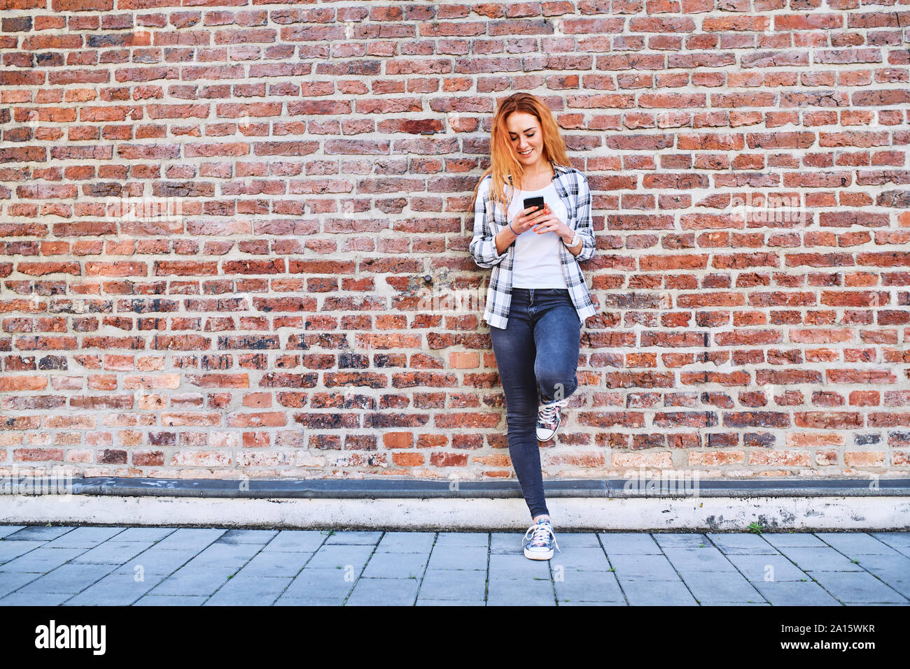 Mujer joven recostada contra la pared de ladrillo en la ciudad mientras utiliza el smartphone Foto de stock