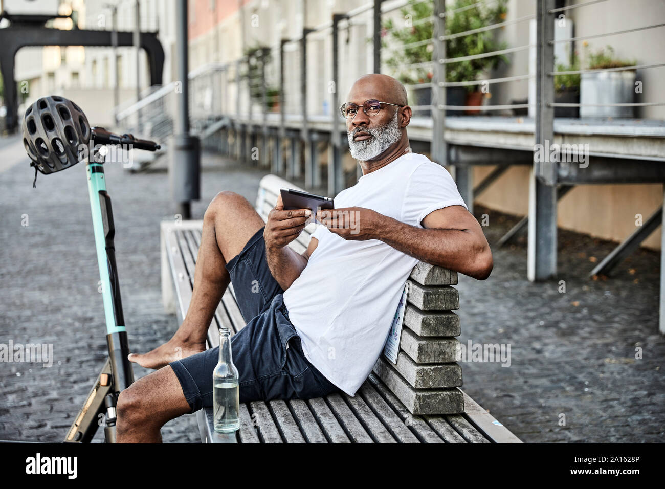 Retrato de hombre maduro con E-Scooter y teléfono celular sentado en un banco en verano Foto de stock