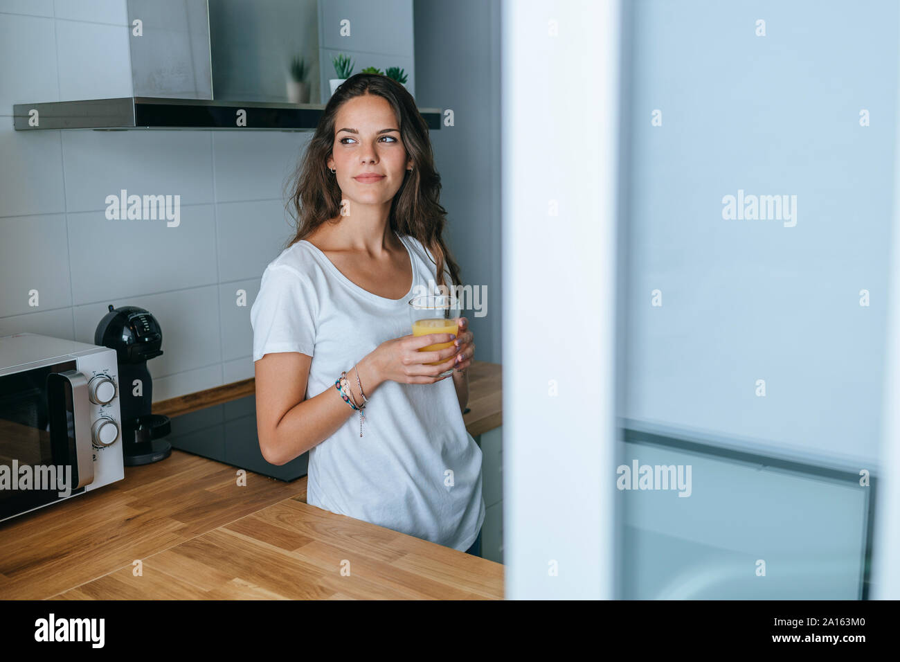 Mujer joven de pie en la cocina con un vaso de jugo de naranja Foto de stock