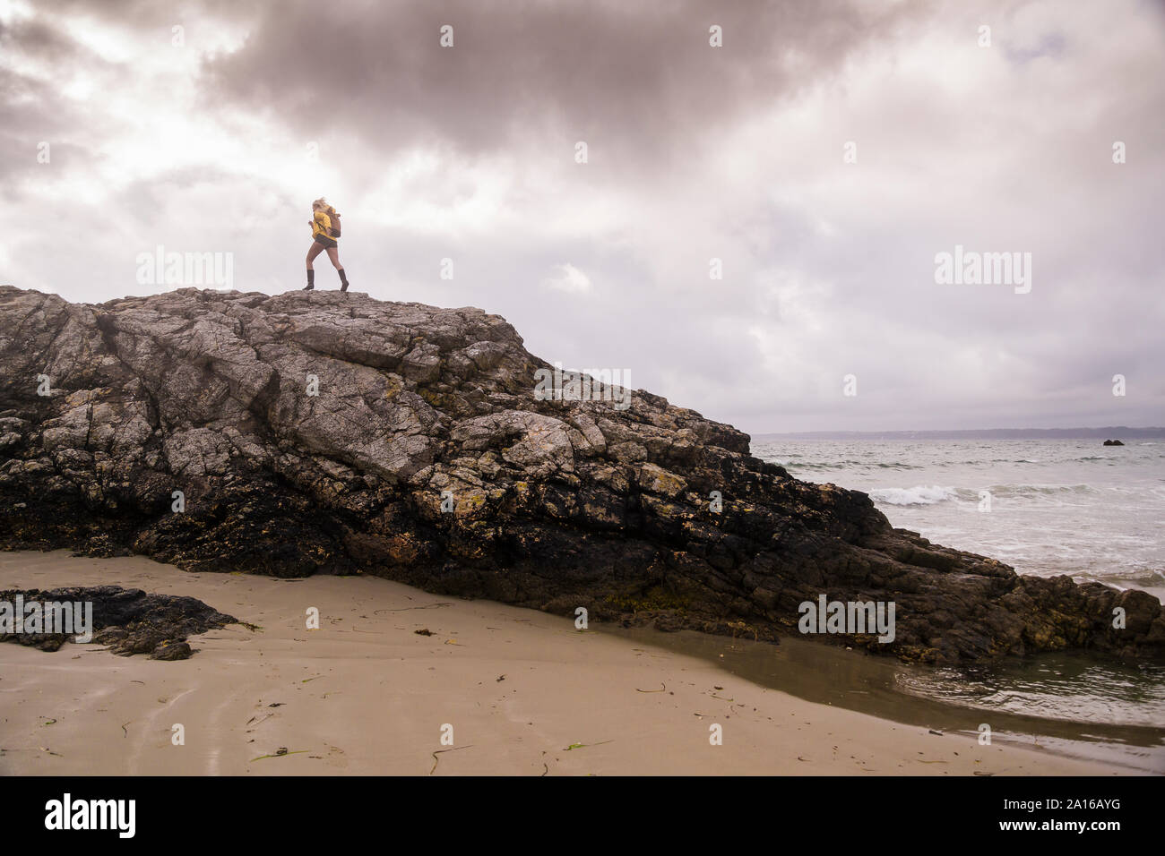 Mujer vistiendo chaqueta de lluvia amarilla de pie en la playa rocosa Foto de stock