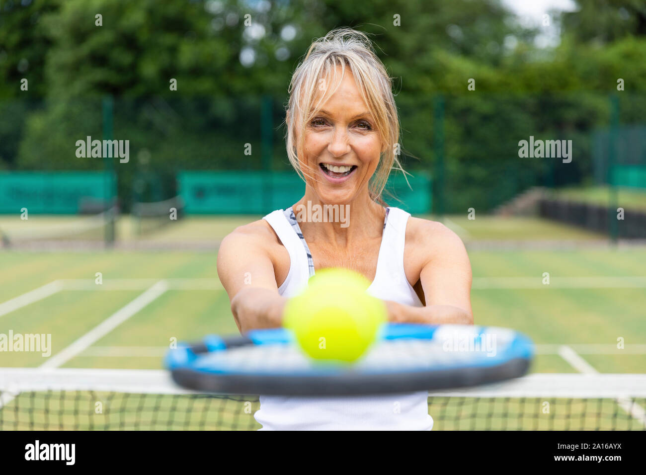 Retrato de mujer madura de risa sosteniendo una raqueta de tenis con una pelota en el club de tenis. Foto de stock