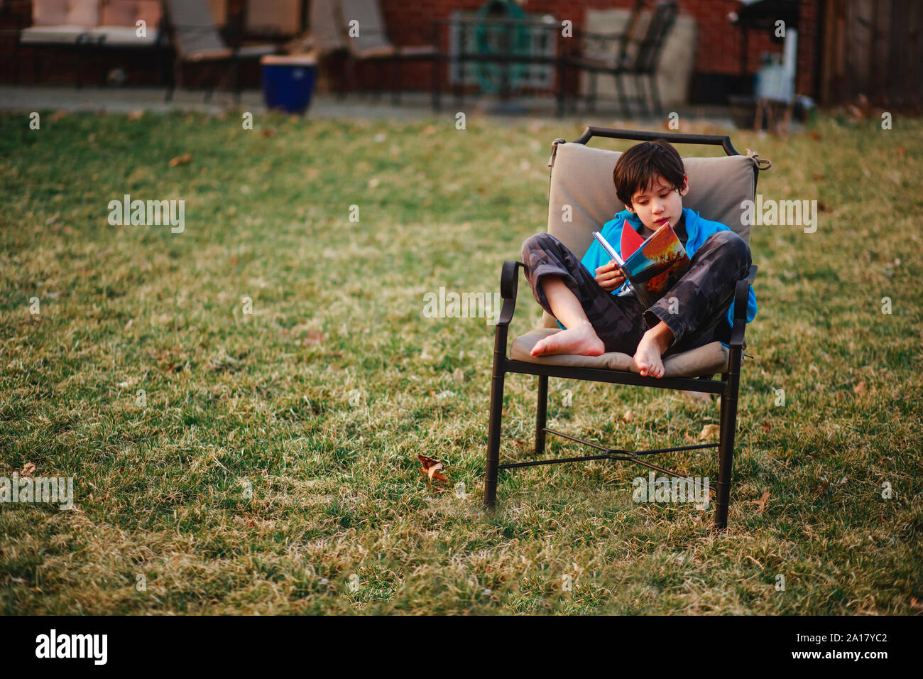 Una niña pequeña está sentada en una silla de pies descalzos en el patio leyendo un libro en la primavera Foto de stock