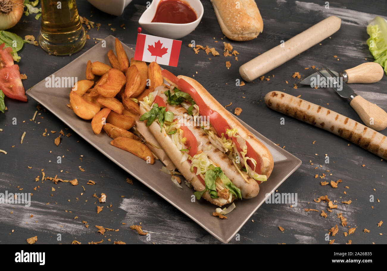 Perro caliente casero con salchichas blancas con diversos condimentos y la bandera Canadiense vistos desde arriba en la tabla de madera gris Foto de stock