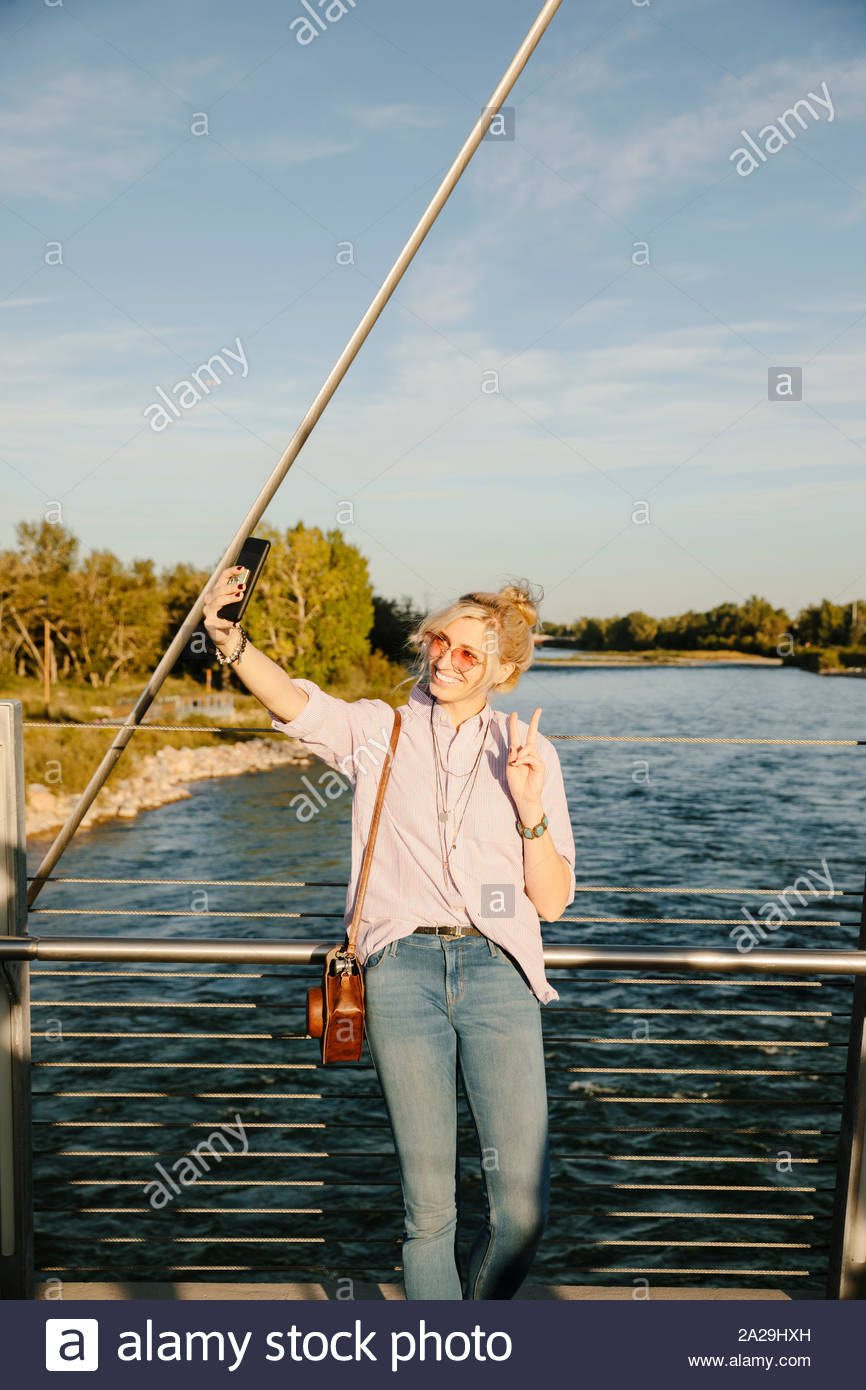Mujer joven tomando selfie teléfono con cámara en el puente con vistas soleado río Foto de stock