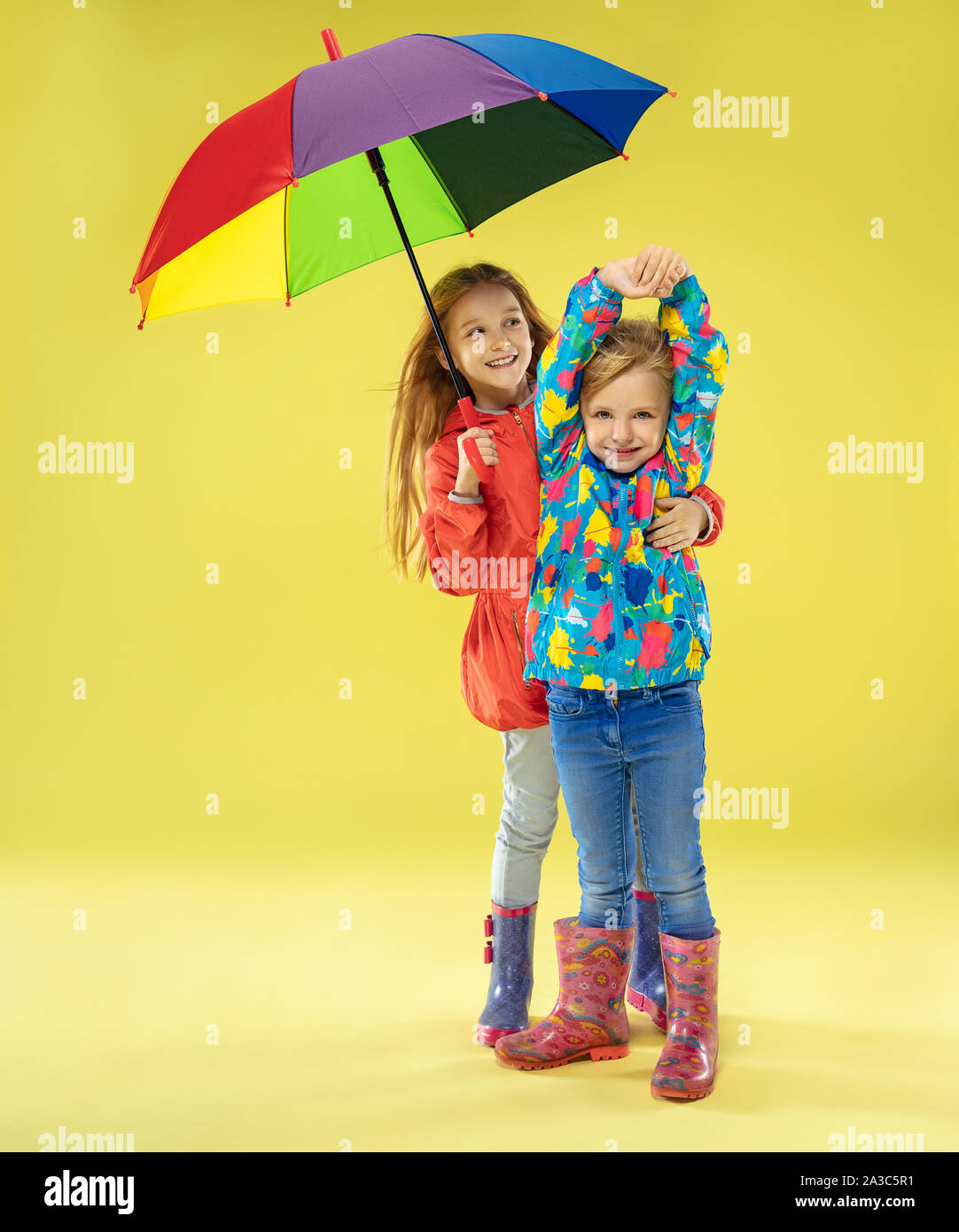 Una longitud completa de un brillante retrato de Moda en un impermeable sosteniendo un paraguas de colores del arco iris sobre fondo amarillo studio. Otoño y primavera moda para niños.