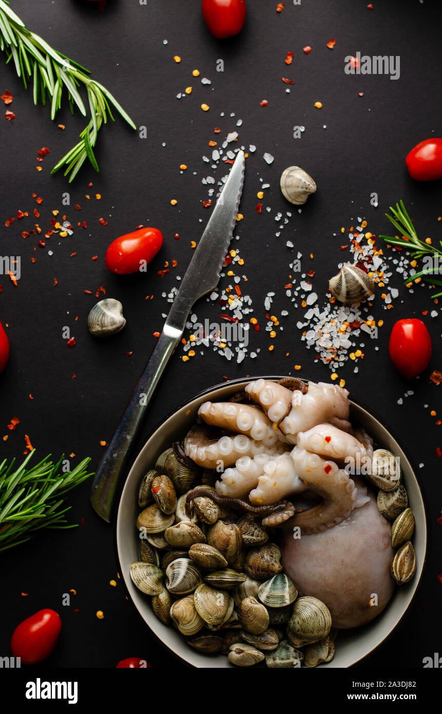 Un recipiente con pulpo fresco con almejas y los ingredientes para cocinar sobre un fondo negro. Concepto de comida mediterránea. Vertical, vista superior Foto de stock