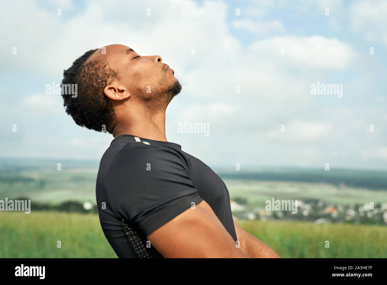 Cerca de guapo, deportivo hombre africano formación y meditar. Deportista vestían de negro camiseta hacer yoga. Atleta muscular posando al aire libre con los ojos cerrados. Foto de stock