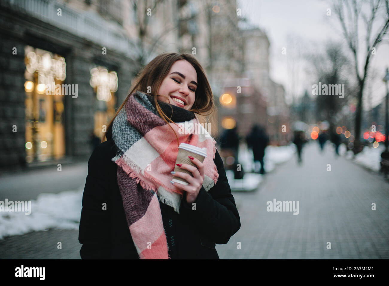 Feliz joven con copa desechable vistiendo ropa de abrigo mientras caminaba por las calles de la ciudad durante el invierno Foto de stock