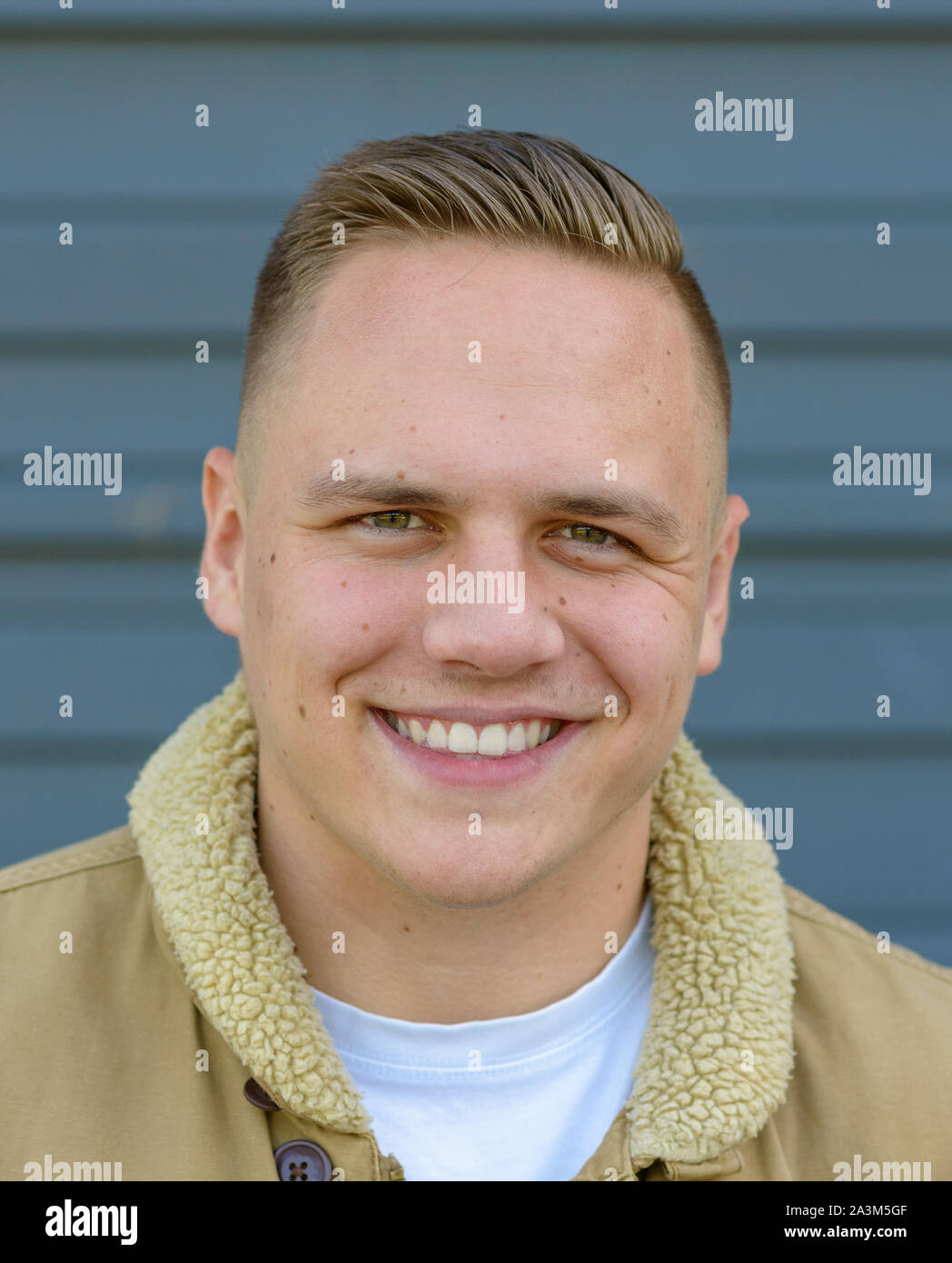 Retrato de un hombre joven con una amplia sonrisa encantadora, mirando la cámara contra una pared gris Foto de stock