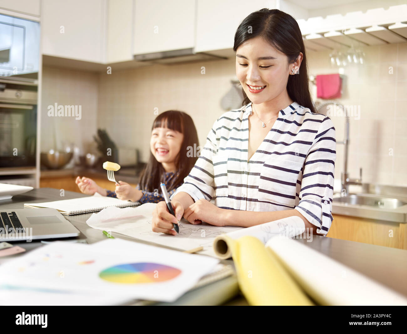 Jóvenes profesionales de diseño asiático madre trabajando en casa teniendo cuidado de la hija. Foto de stock