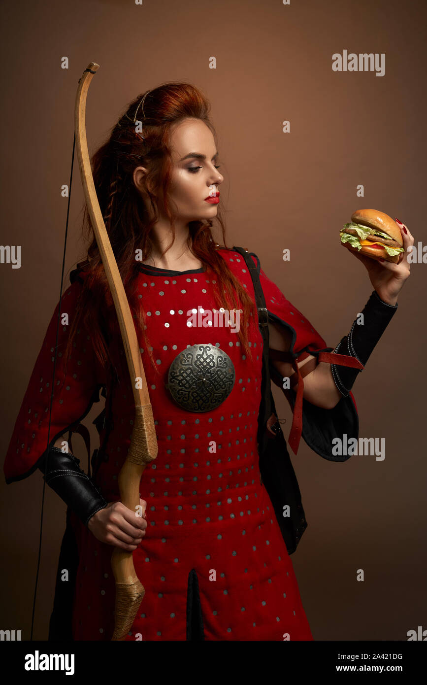 Vista desde el lado del pelo rojo hembra guerrero con arco y flecha manteniendo sabrosas hamburguesas sobre antecedentes aislados en el estudio. Determina el hambre héroe femenino comer comida rápida. Concepto de poder. Foto de stock