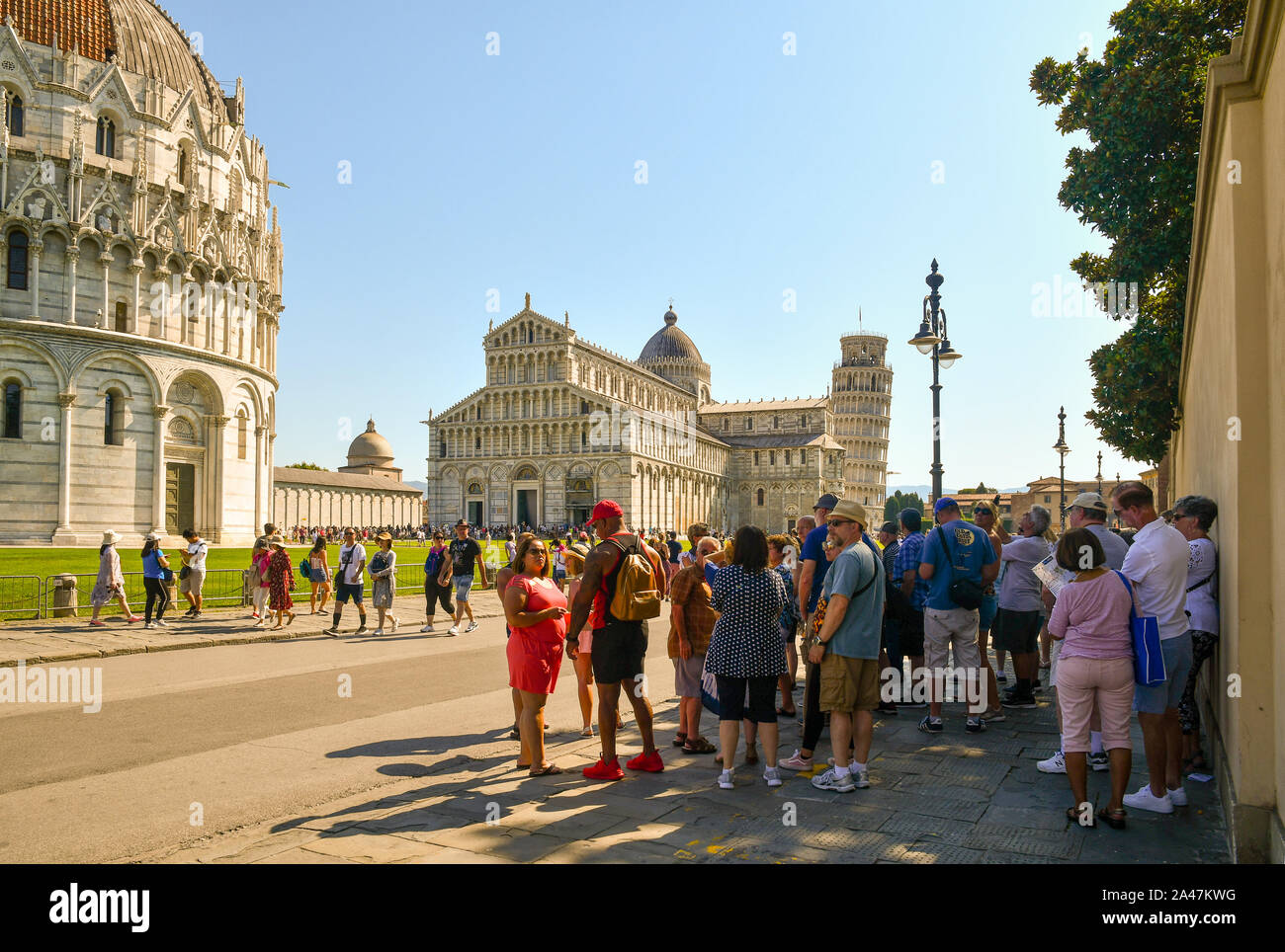 Un grupo de turistas descansando en la sombra de un muro en la famosa Piazza dei Miracoli con la torre inclinada en un caluroso día de agosto, Pisa, Toscana, Italia Foto de stock