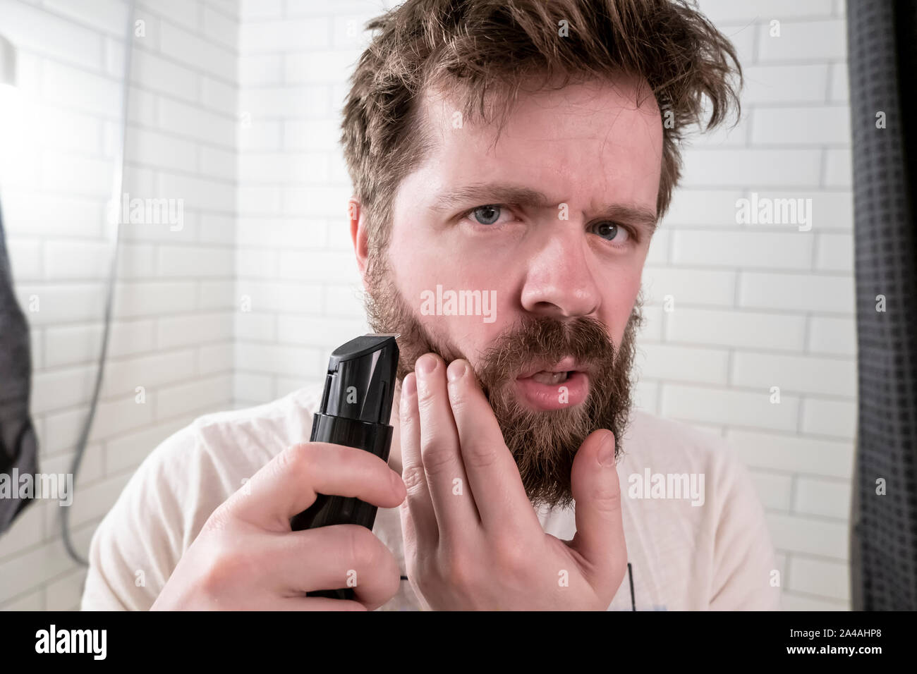 Malestar joven mira en el espejo a su shaggy peinado y espesa barba y va a  hacerse un corte de pelo con la ayuda de una fresa. Clos Fotografía de  stock -