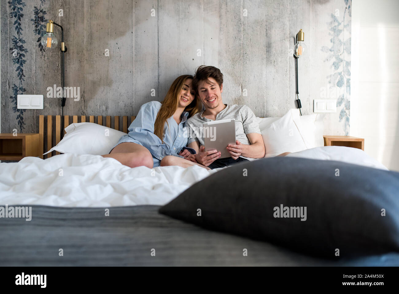Feliz pareja joven caricias acostada en la cama en casa utilizando un tablet Foto de stock