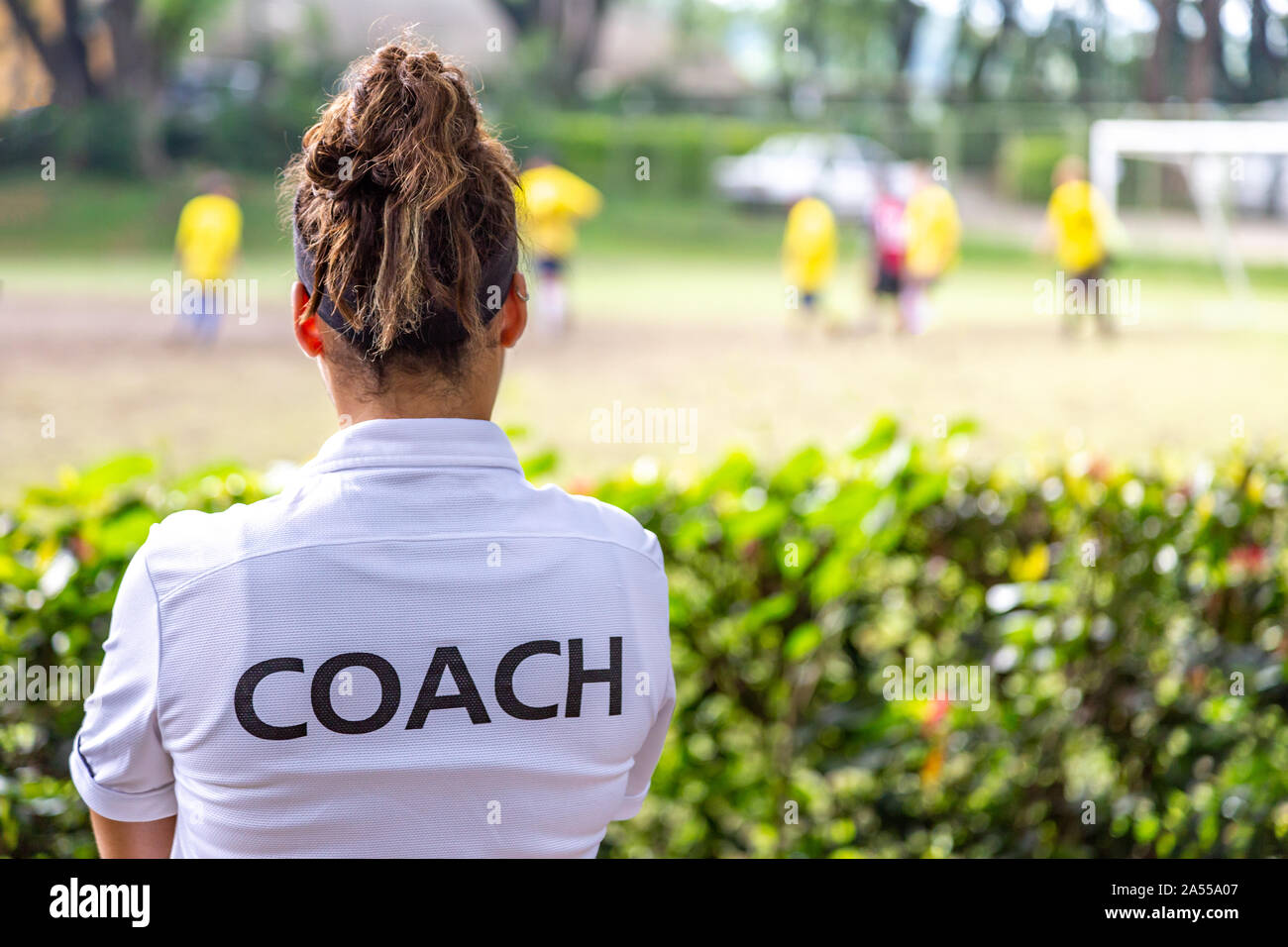 Vista posterior de un fútbol femenino, fútbol, entrenador en entrenador blanco Camiseta viendo a su equipo jugar en un campo de fútbol al aire libre Foto de stock