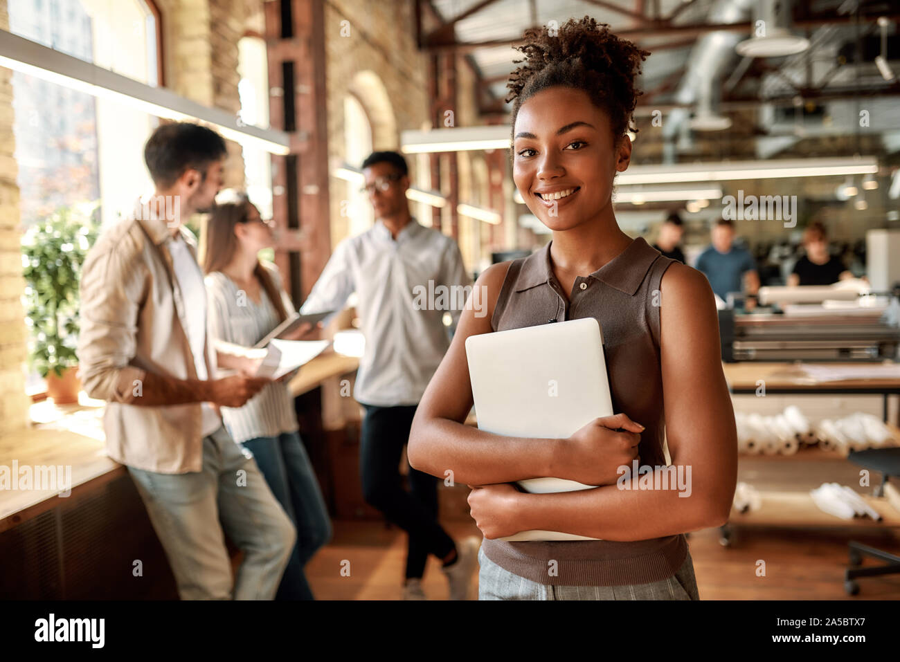 Feliz de trabajar aquí. Alegre y joven afro americana mujer sosteniendo portátil y sonriendo mientras está de pie en la oficina moderna. Foto de stock