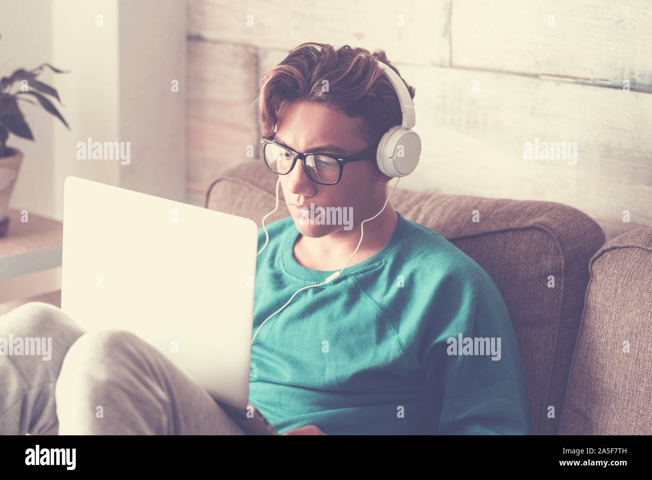 Joven estudiante en casa con un moderno equipo portátil auriculares y estudiar con internet cnnection - guapo muchacho sentado en el sofá con tecnología Foto de stock