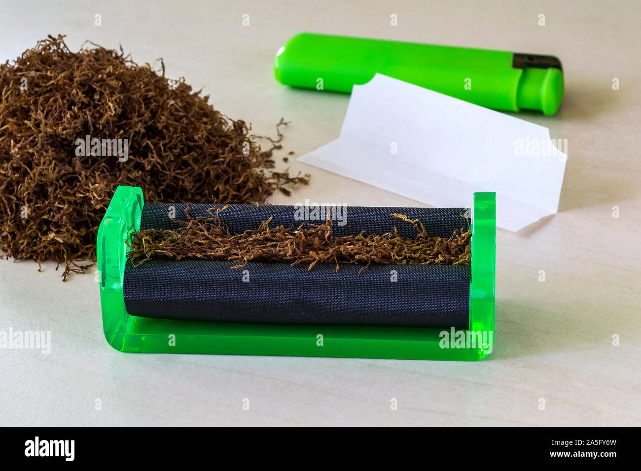 Cigarrillo verde máquina laminadora, papel de cigarrillo, verde ligero y el montón de tabaco en una tabla en blanco. Hacer los cigarrillos con tabaco de pipa en casa. Foto de stock