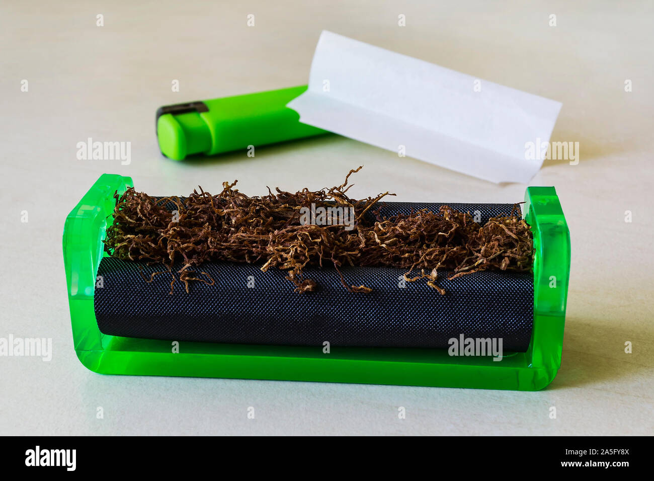 Verde máquina laminadora de cigarrillos con tabaco, papel de fumar y encendedor verde sobre una tabla en blanco. Hacer los cigarrillos con tabaco de pipa en casa. Foto de stock
