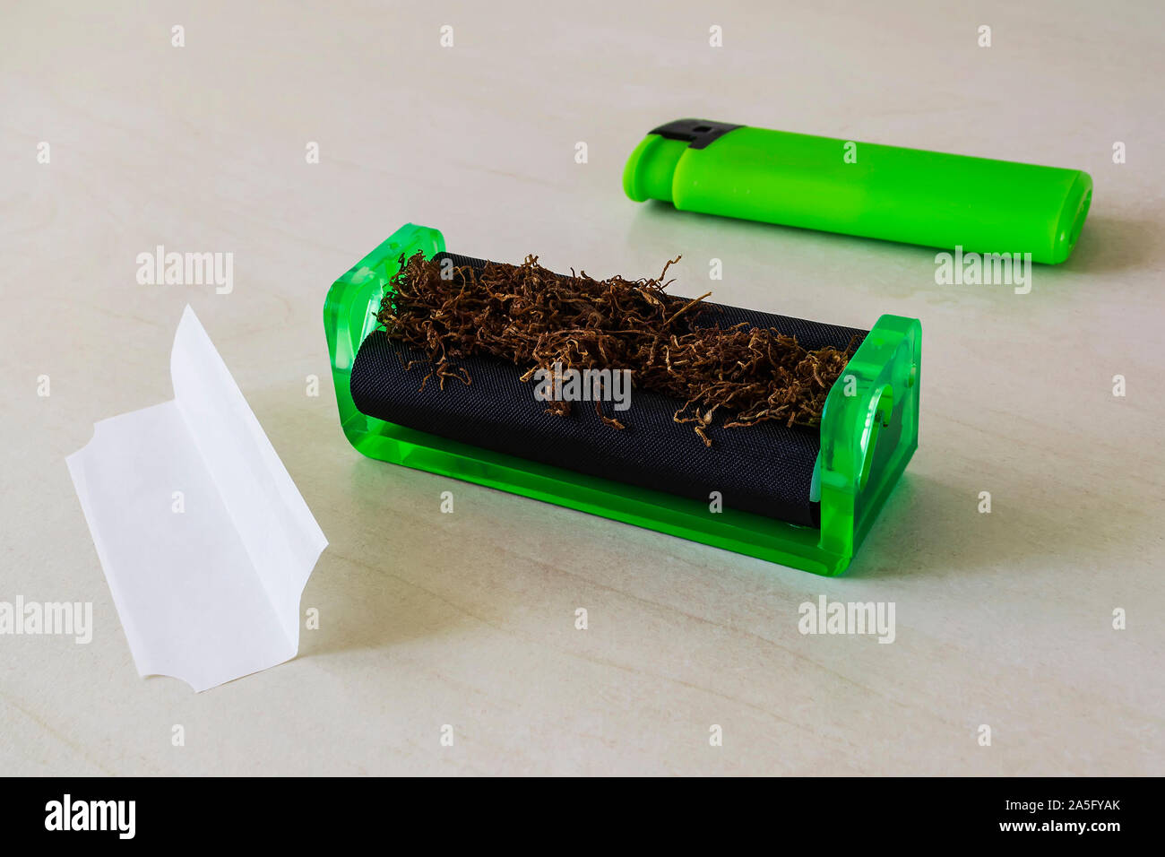 Verde máquina laminadora de cigarrillos con tabaco, verde ligero y papel de cigarrillo sobre una tabla en blanco. Hacer los cigarrillos con tabaco de pipa en casa. Foto de stock