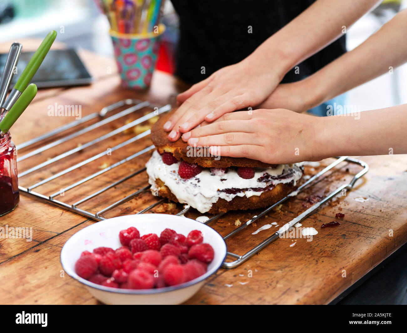 Chica y su hermana hornear un pastel, presionando hacia abajo la capa superior en la mesa de la cocina, vista recortada de manos Foto de stock