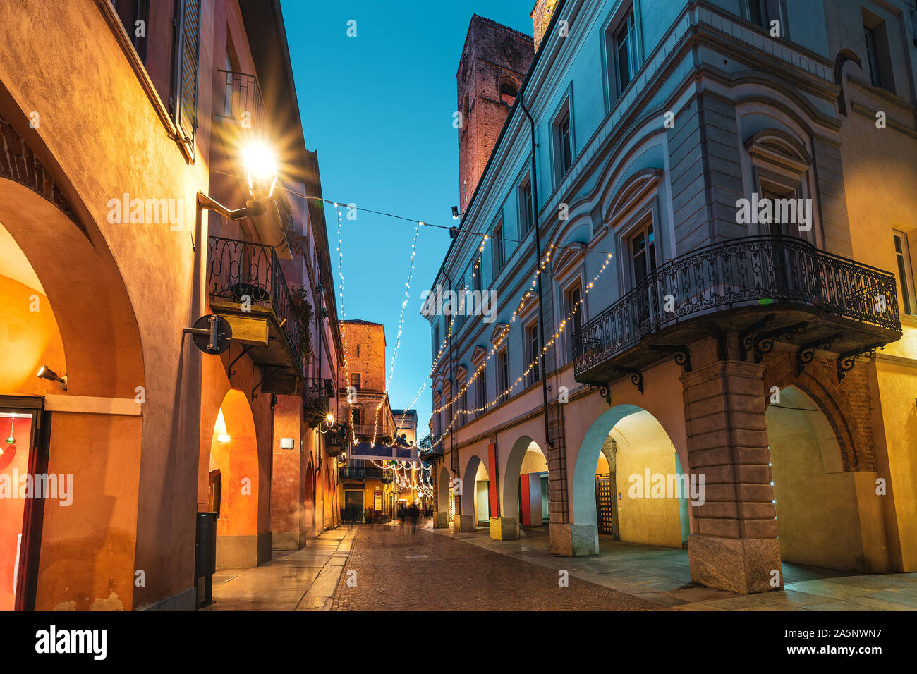 Calle peatonal adoquinada iluminada de noche en el casco antiguo de la ciudad de Alba, Piamonte, Norte de Italia. Foto de stock