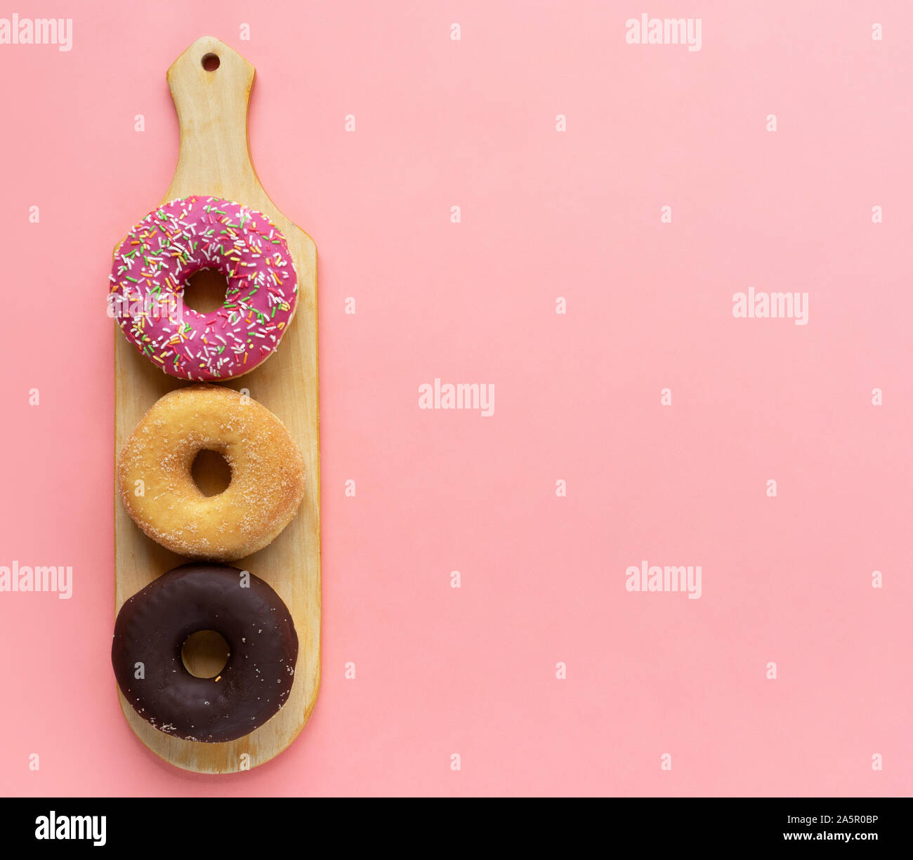 Tres diferentes donuts, chocolate donut, Rosa guinda y sin guinda sobre una tabla de cortar de madera sobre un fondo de color rosa Foto de stock