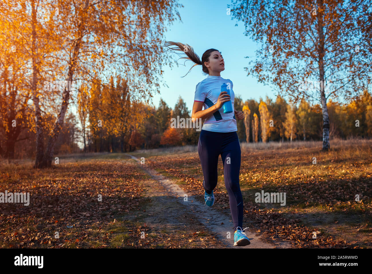 Runner formación en otoño de parque. Mujer corriendo con la botella de agua y mantenerse en forma en la puesta de sol. Un estilo de vida activo. Entrenamiento Foto de stock