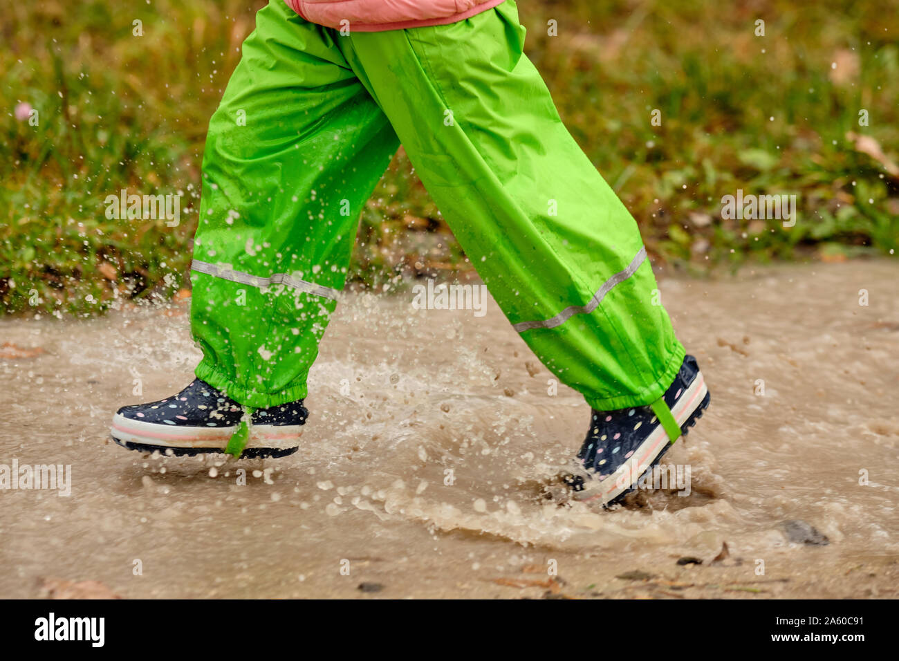 Vista lateral de la sección baja de un niño niña en verde pantalón  impermeable y botas de goma que se ejecutan a través de un gran charco de  lluvia en el bosque