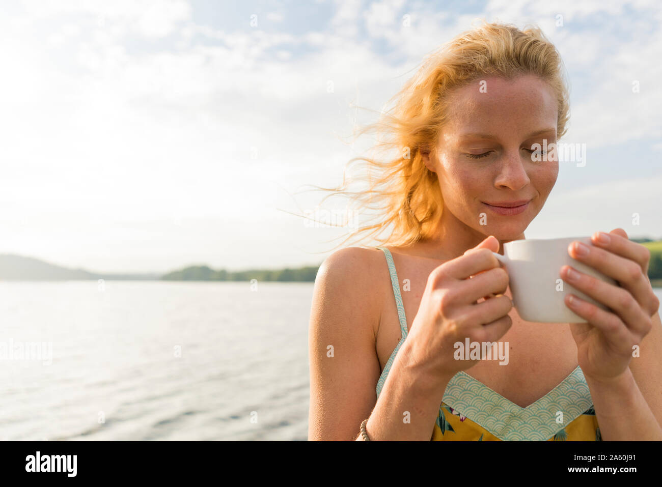 Mujer joven con los ojos cerrados dwith sosteniendo una taza en un lago Foto de stock