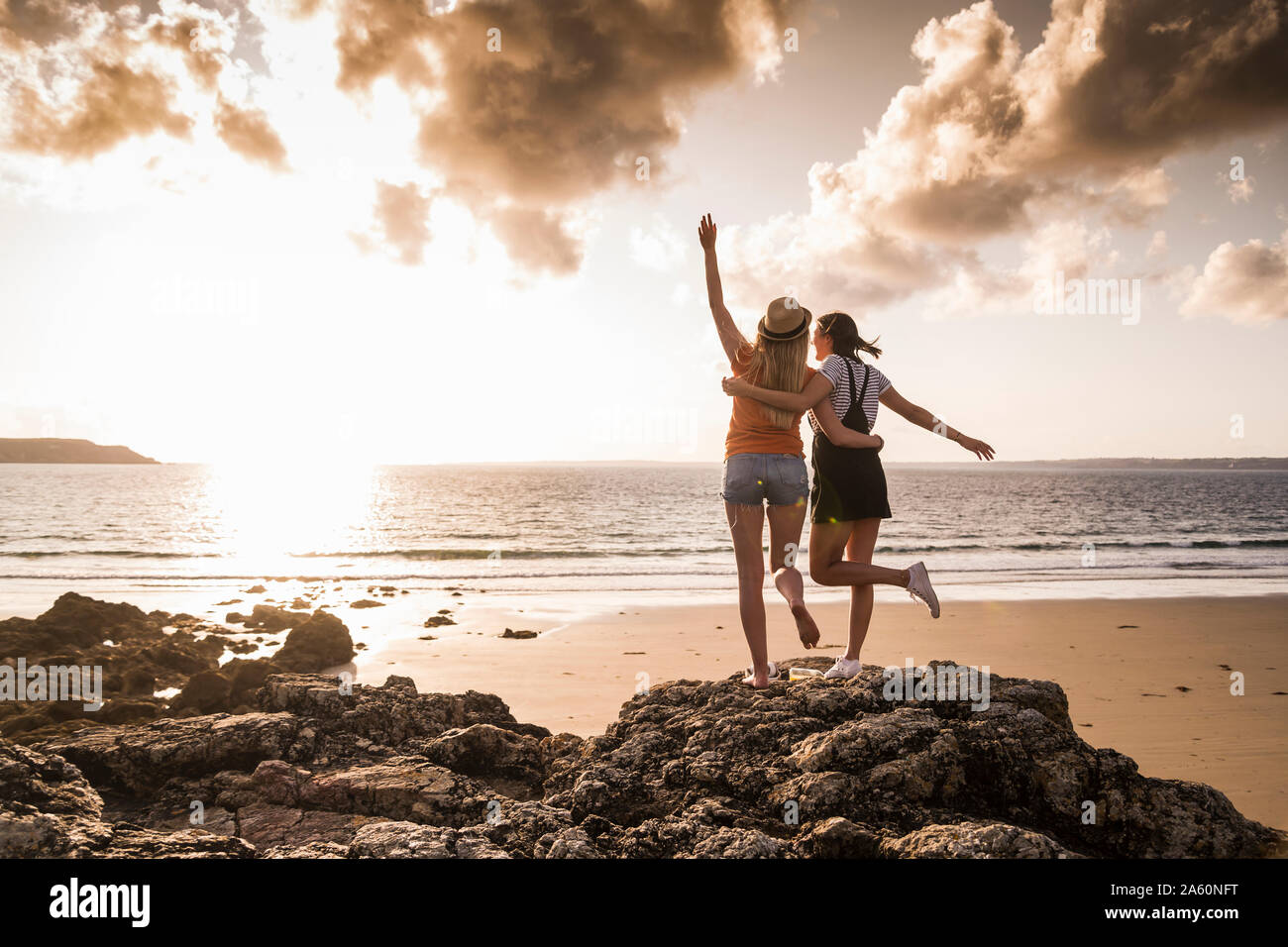 Dos amigas de pie en la playa rocosa, saludando al atardecer, vista trasera Foto de stock
