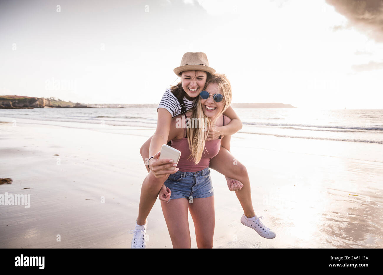 Dos amigas divirtiéndose en la playa, llevando mutuamente, teniendo piggyback selfies smartphone Foto de stock