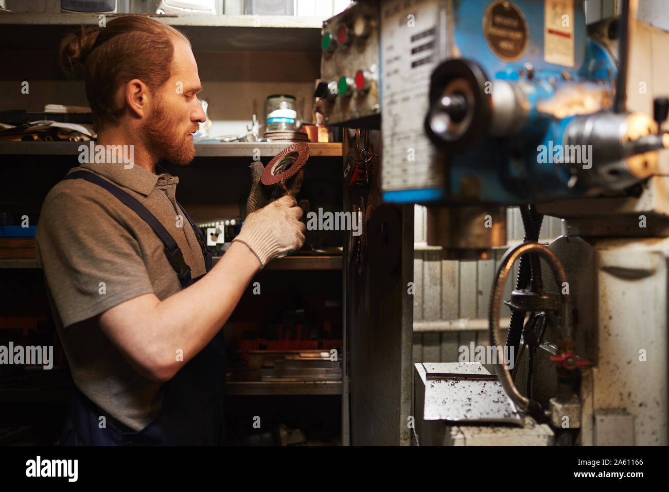 Joven barbudo mecánico examinar detalles de metal mientras trabajaba en el almacén Foto de stock