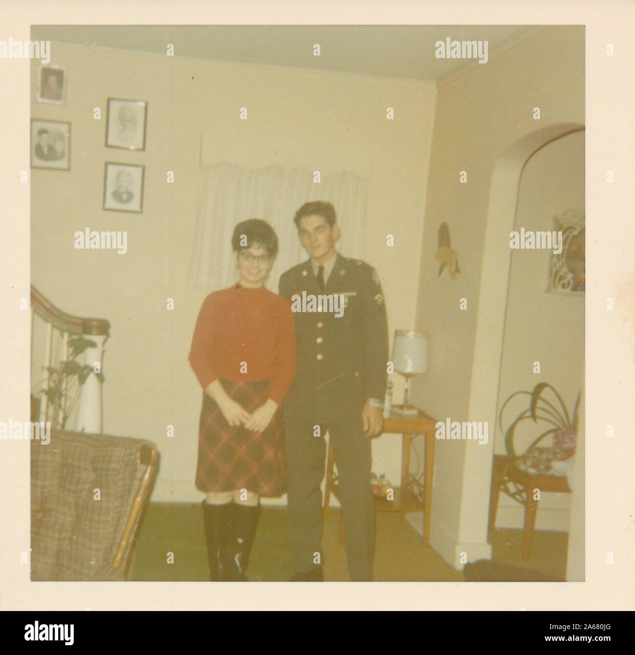 Disparo completo de un militar uniformado posando con una joven sonriente, ambos frente a la cámara, mientras se encuentra cerca de la base de una escalera en una sala de estar suburbana, Estados Unidos de América, 1965. () Foto de stock