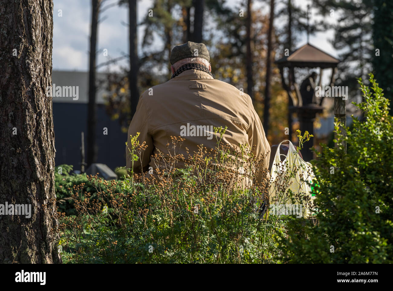 Un viejo hombre gris en una capa ligera y hat llora por su fallecimiento tumba en el cementerio de la caída en un día soleado. Una cesta colocada junto a un hombre. Foto de stock