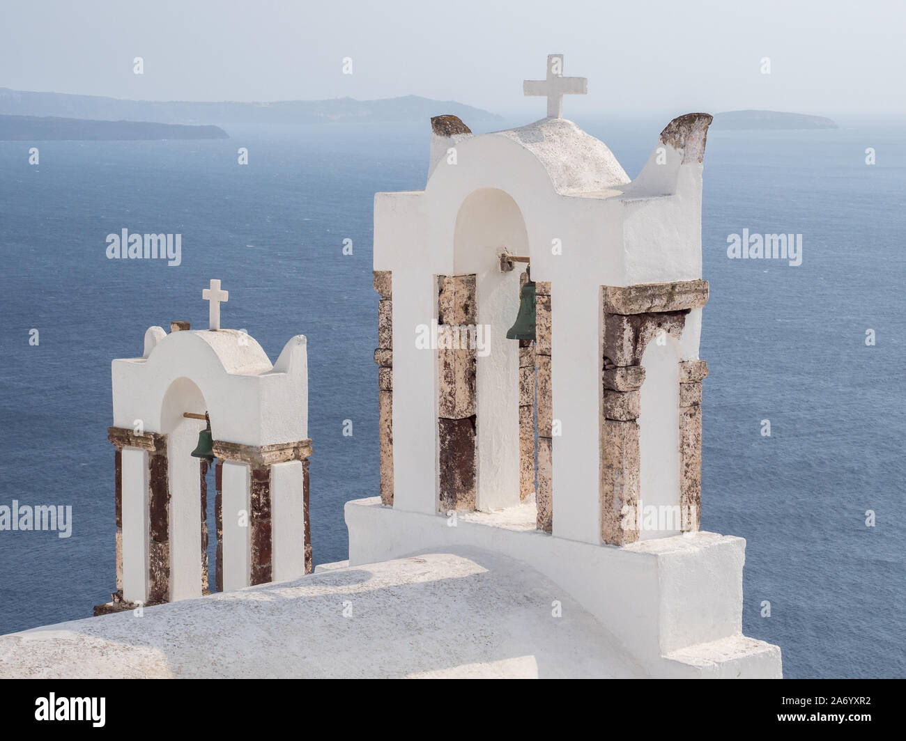Un par de color blanco lavado dos torres con campanario cruz crucifijo cruza el mar Mediterráneo y las montañas de fondo agua horizonte Oia Santorini Foto de stock