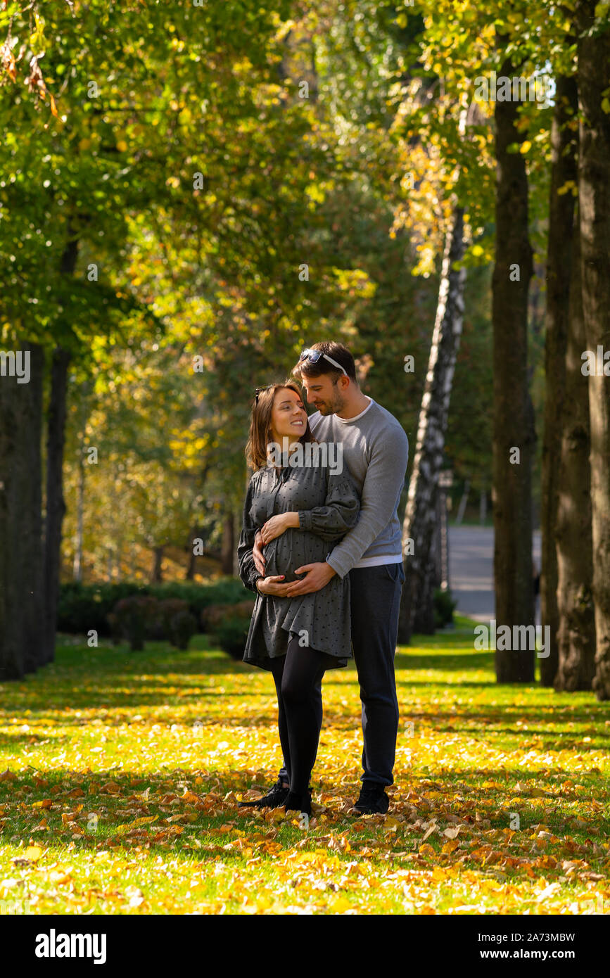 Pareja joven con la esposa embarazada de pie en un parque en otoño en un abrazo acunando a su estómago, mientras que el vínculo con su hijo por nacer Foto de stock