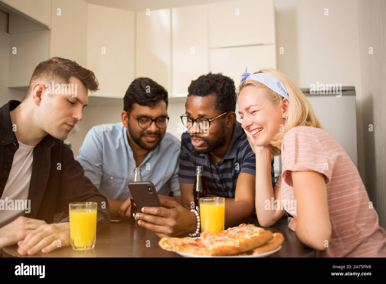 Cuatro amigos sentados a la mesa, comer pizza, beber bebidas gaseosas, la lectura de un mensaje de texto en el teléfono inteligente, prueba nueva aplicación teléfono juntos. Mult Foto de stock