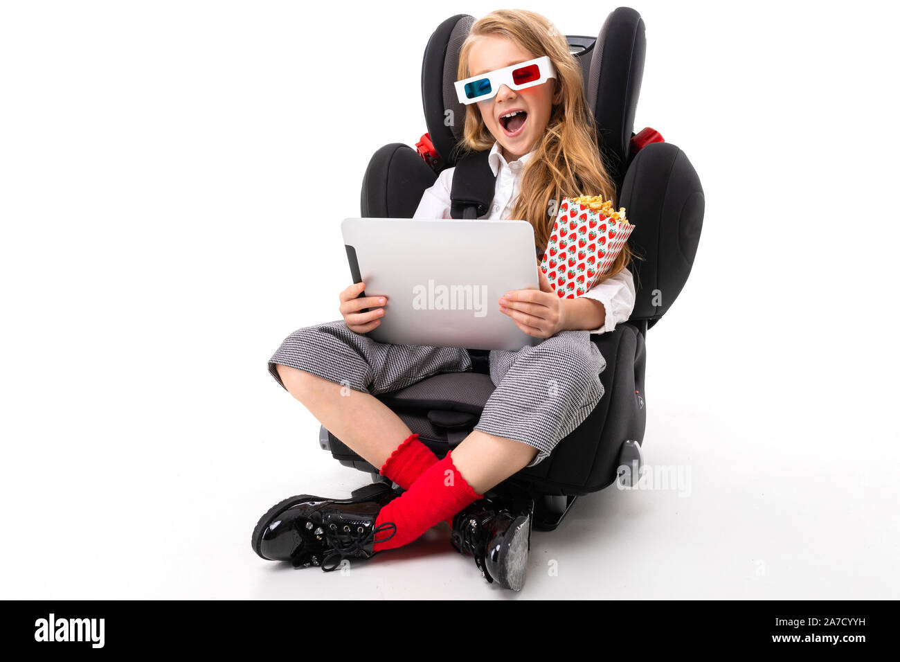 Una niña con maquillaje y largo pelo rubio sentado en una silla de  automóvil para bebés con tablet, auriculares, pop-corn, Gafas 3D y ver la  interesante película de Fotografía de stock -