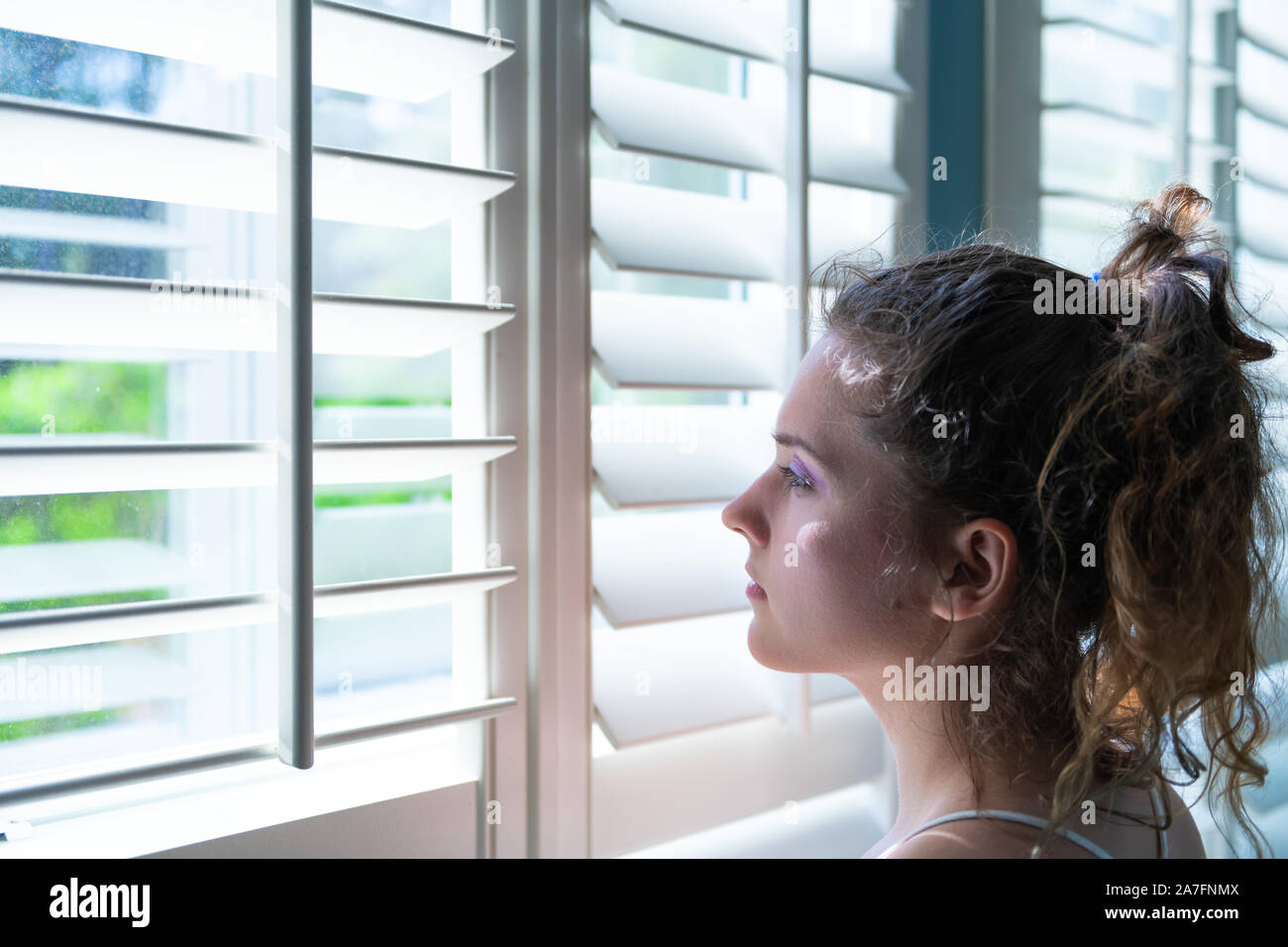 Una joven mujer de pie mirando a través del vidrio blanco persianas de ventana en la habitación de la casa, casa o apartamento en persianas la luz del de