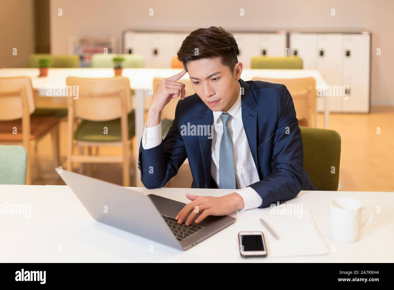 Cintura para arriba la foto de un joven empresario con pelo negro y un traje sentado concentrado en trabajar en la parte frontal del portátil en el escritorio en una oficina. Foto de stock