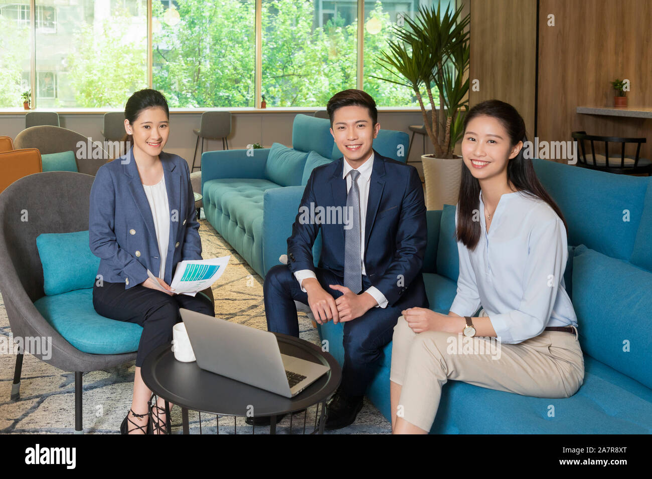 Retrato de tres empresarios macho y hembra sonriente sentado en una oficina con un portátil sobre la mesa Foto de stock