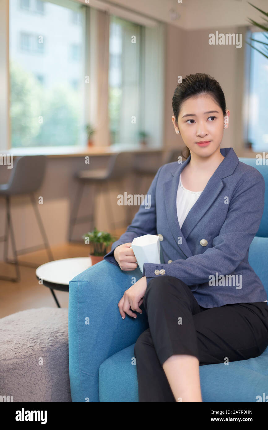 Retrato de una joven empresaria con pelo negro sentado en un sillón azul en una oficina y sosteniendo la taza con café Foto de stock