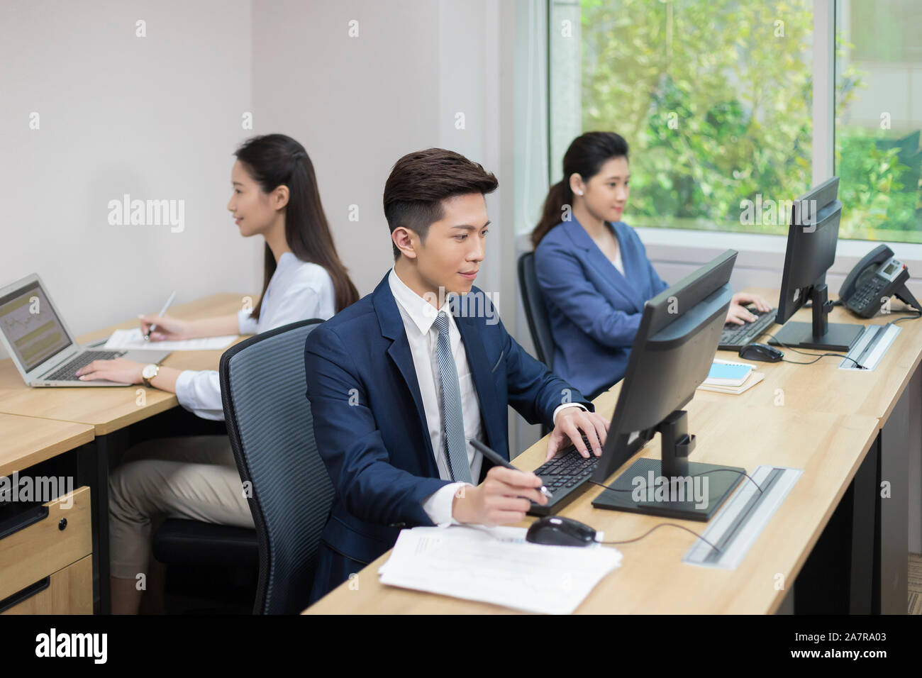 Fotografía de tres empresarios jóvenes, hombres y mujeres que trabajan en una oficina con dos usando computadoras y una utilización portátil Foto de stock