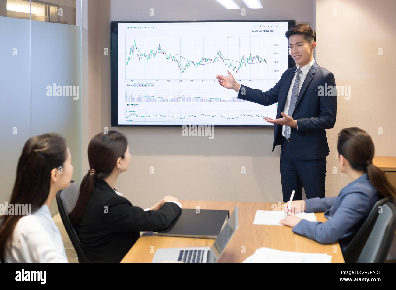 Joven hombre de negocios sonriente en un traje dando una presentación en una sala de juntas con un gráfico en una pantalla detrás de él y con otros tres empresarios Foto de stock
