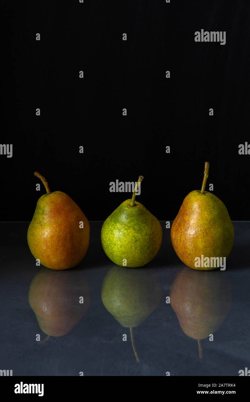 3 peras maduras de amarillo, verde y rojo color gris acostarse sobre una mesa brillante sobre un fondo negro. Las peras se refleja en la superficie de la mesa. Foto de stock