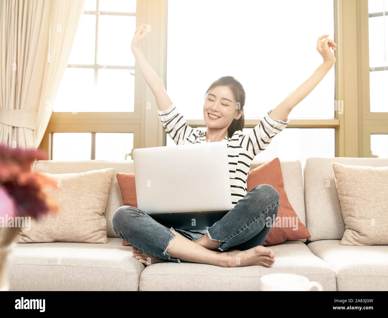 Hermosa y alegre joven mujer asiática trabaja desde casa sittng en sofá con ordenador portátil, con los brazos en alto celebrando el éxito Foto de stock