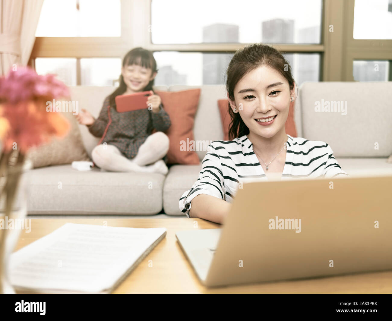 Joven mujer asiática madre trabajando en casa utilizando equipo portátil con mi hija en segundo plano. Foto de stock