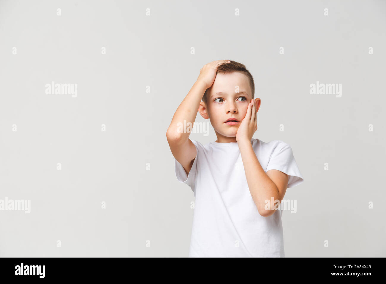 Asustado muchacho en camiseta blanca con ambas manos sobre la cabeza sobre fondo gris Foto de stock