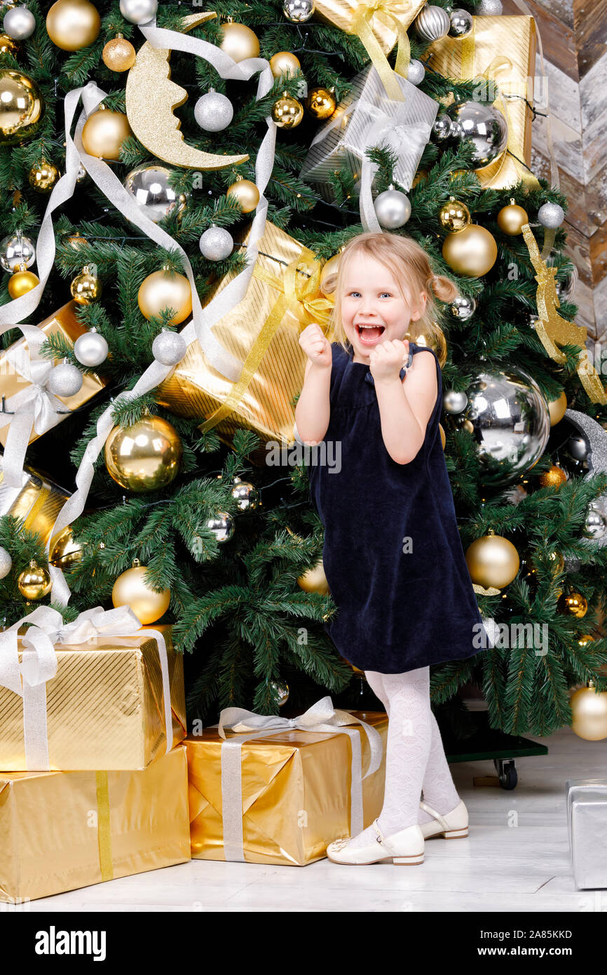 Retrato de gozosa vestida de edad primaria vestido azul stanging junto al árbol de navidad feliz para conseguir regalos de Navidad Foto de stock